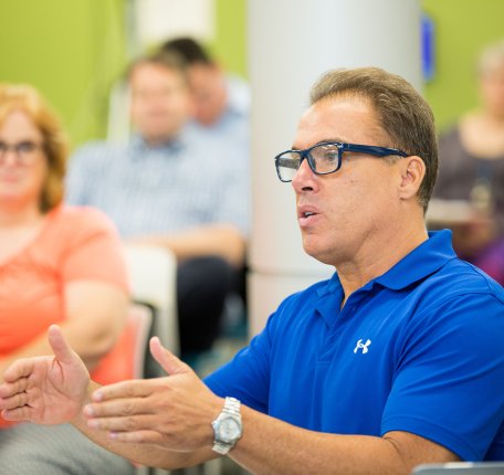 A male instructor in a blue polo shirt and glasses speaking animatedly during a classroom or meeting discussion, with other participants seated in the background.