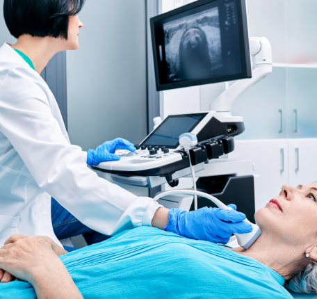A female clinician in a white coat and blue gloves performing an ultrasound on a patient's neck, with the sonogram displayed on a monitor above.